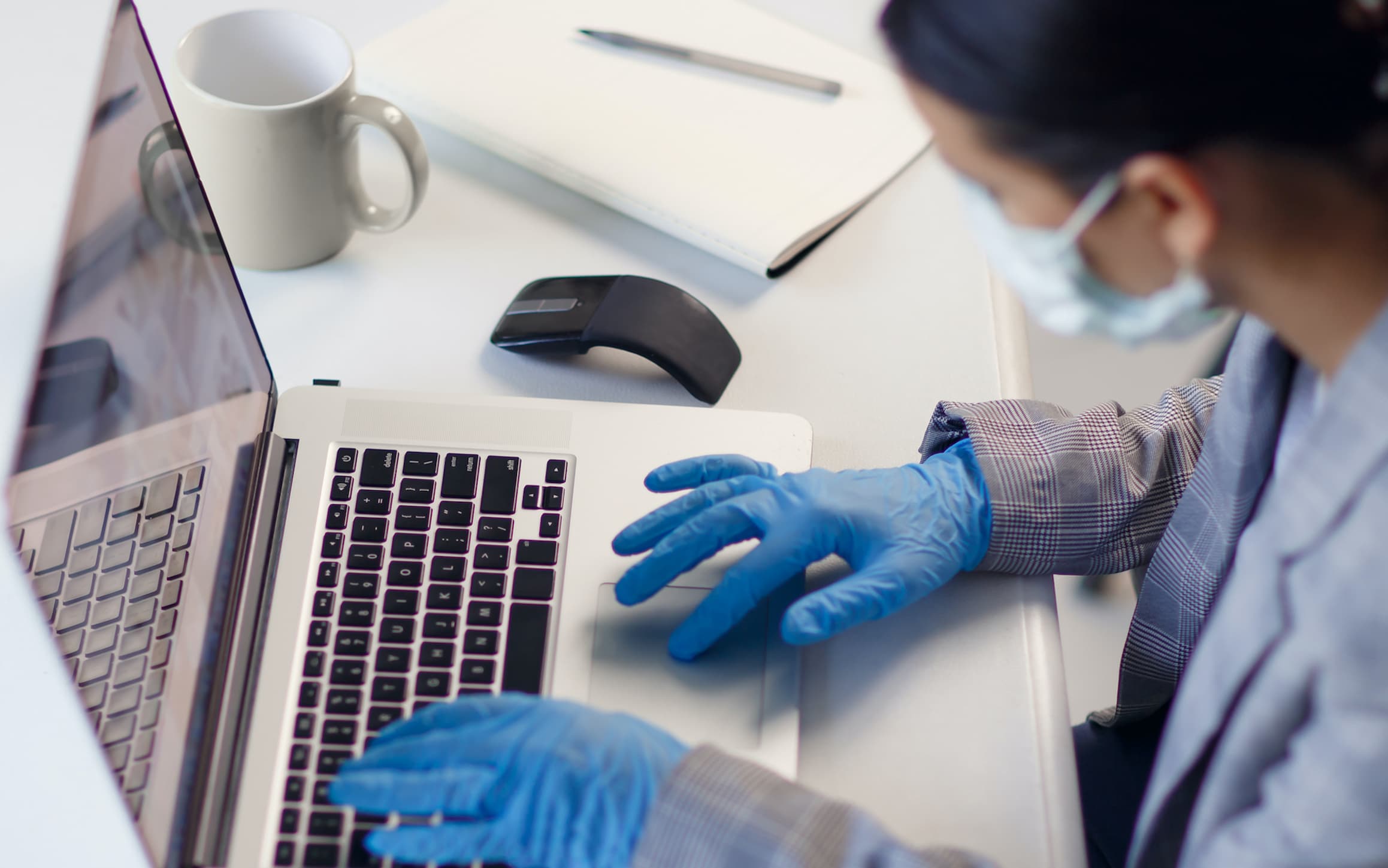 Young woman employee wearing protective gloves on hands and mask on face working from office or home using laptop on desk. Preventing corona virus covid-19 spread during epidemic outbreak